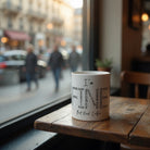 White ceramic mug with text describing various feelings, sitting on a wooden table by a window.