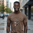 Brown unisex cotton t-shirt featuring a distressed print of a raccoon holding a coffee cup.