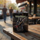 Black ceramic mug with a Grim Reaper tailor graphic holding a seam ripper on a wooden outdoor table.