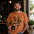 Man in an orange t-shirt with a squirrel graphic and the text Feral For Coffee holding a black mug.