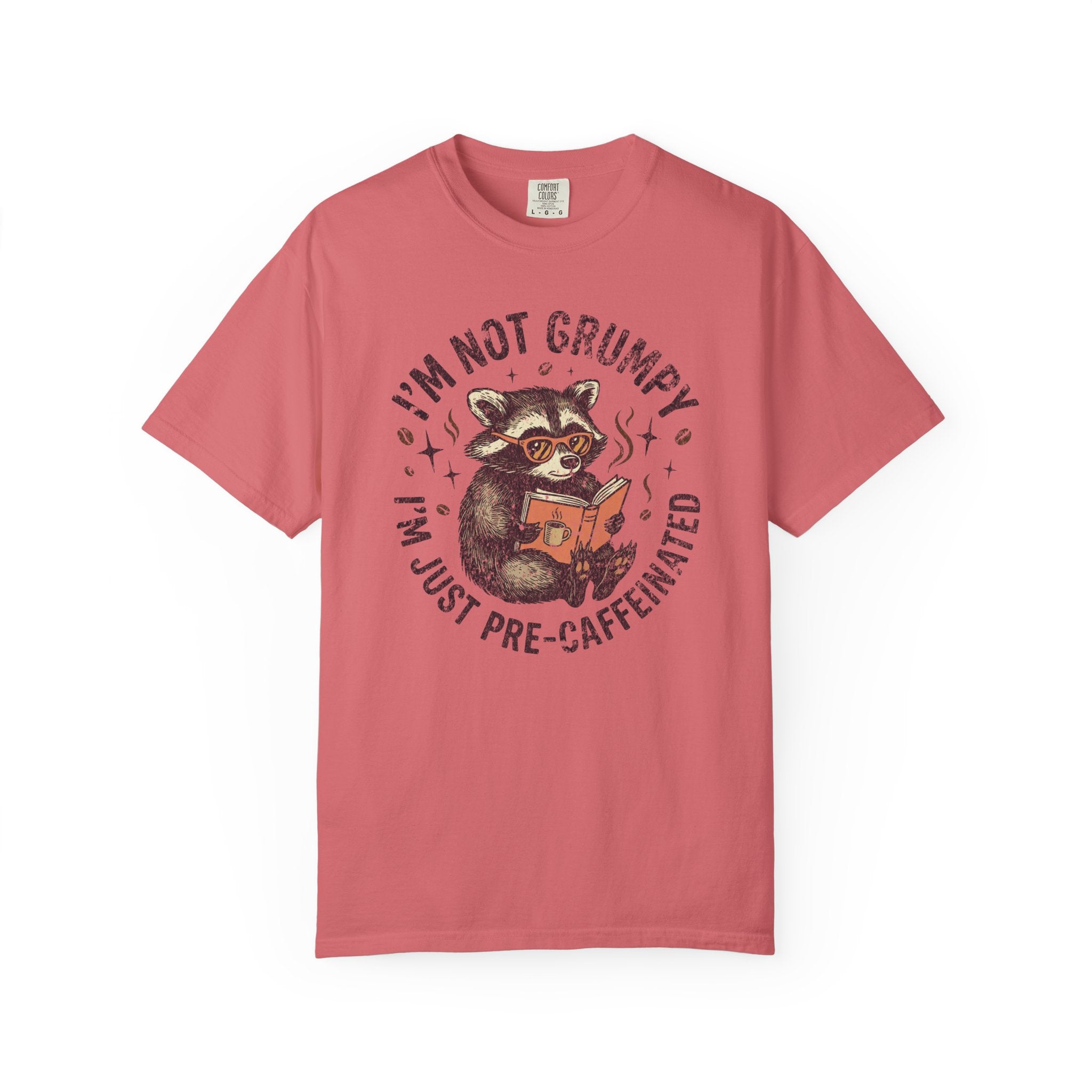 Coral-colored unisex t-shirt featuring a graphic of a raccoon reading a book with coffee.