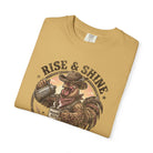 Folded tan t-shirt featuring a graphic of a cowboy rooster pouring coffee, labeled Rise & Shine.