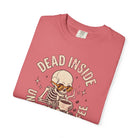 A folded pink t-shirt featuring a distressed graphic of a skeleton wearing sunglasses drinking coffee.