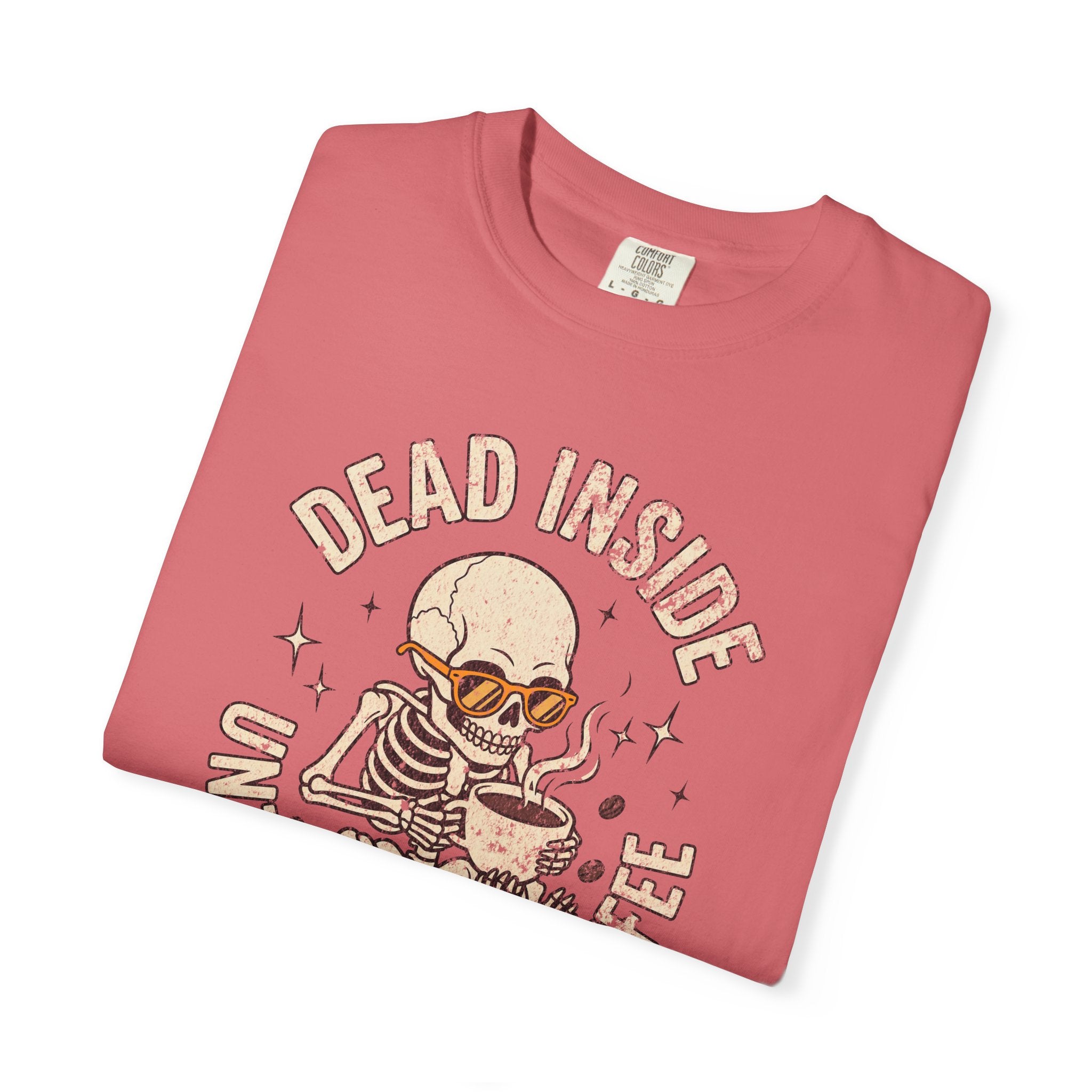 A folded pink t-shirt featuring a distressed graphic of a skeleton wearing sunglasses drinking coffee.