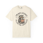 Cream-colored unisex t-shirt featuring a graphic of a raccoon reading a book with coffee.